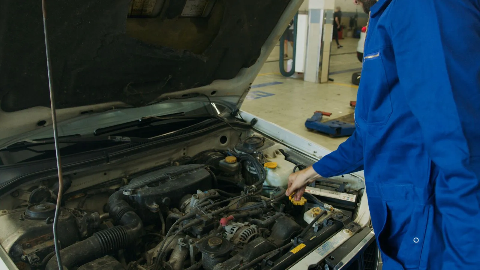 Mechanic in blue coveralls inspecting an open car engine in an automotive workshop.